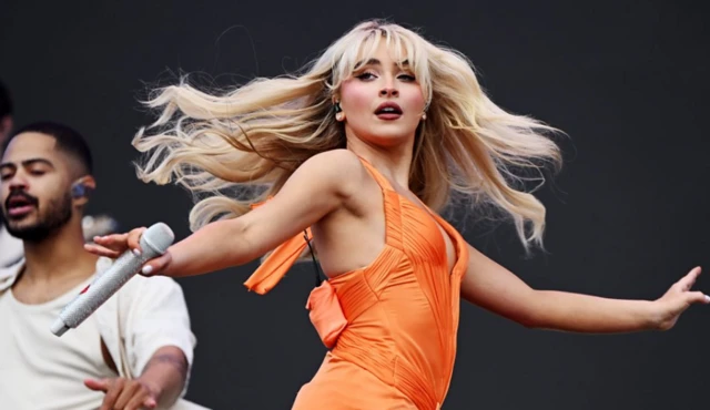 Sabrina Carpenter dancing on stage, wearing orange dress, with microphone in hand.