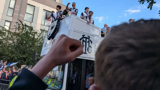 James puts a fist in the air as he looks on at the Tottenham players on top of a white bus.