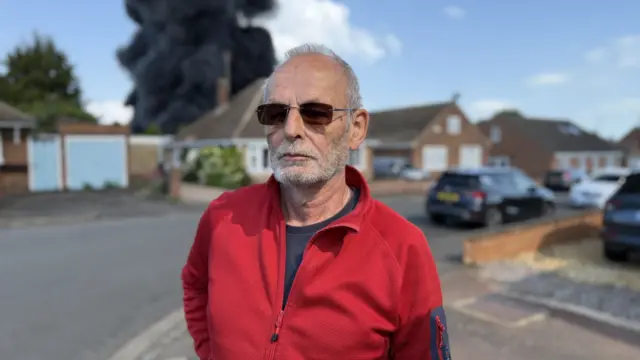 Malcolm stands with dark sunglasses in a squarish frame. He is wearing a red fleece and in the soft focus background smoke from the fire can be seen rising behind residential properties.