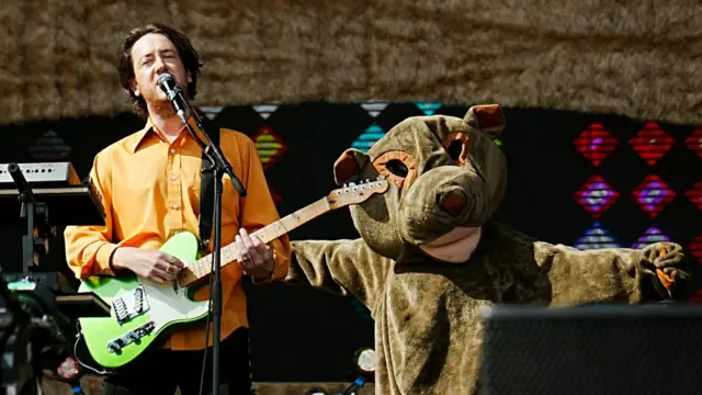 Matthew Murphy of The Wombats sings while holding a green electric guitar. A dancer dressed as a wombat is next to him on stage