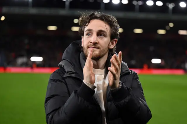 A young man aged around 30 stands on a football pitch applauding. He has dark curly hair and is wearing a black coat.
