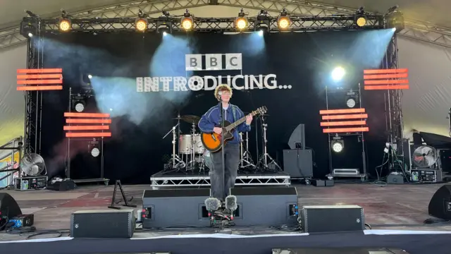 Miles Temp stands on the BBC Introducing Stage with a guitar. He is wearing a navy jacket and black jeans. He is stood in front of a black backdrop that says BBC Introducing.
