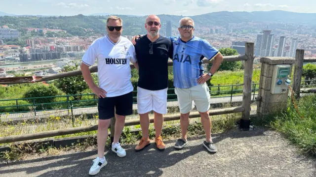 Three men stood arm in arm with the Bilbao skyline behind them