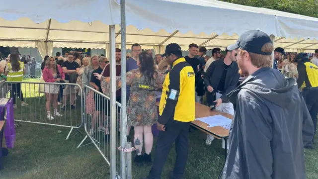 People stand in a queue under a white canopy while security officials carries out checks.