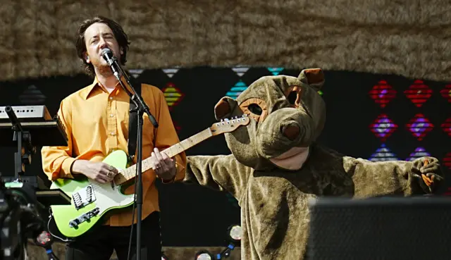 Matthew Murphy plays a green guitar while singing into a microphone. A dancer in a wombat outfit is stood behind him.