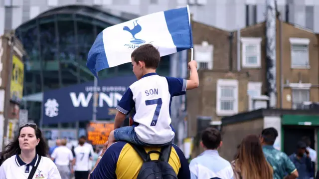 A young boy is pictured on a man's shoulders from behind. He is waving the Spurs flag as they head in the direction of the stadium.
