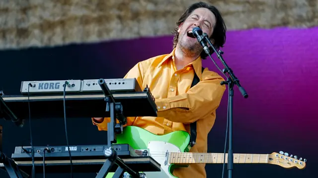 Matthew Murphy sings into a microphone. He has brown hair and is wearing a yellow shirt.