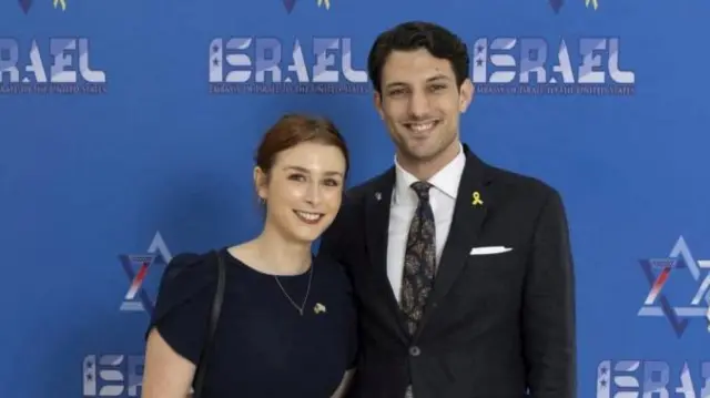 A woman and man stood in front of a blue background with the word Israel printed on it several times. They smile at the camera, as they have one arm around each other looking on