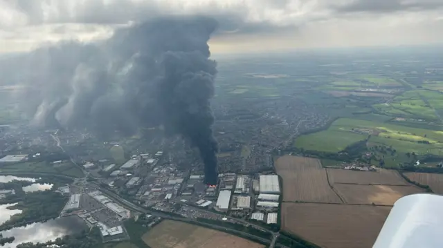 An aerial view of the fire, taken from an airplane, showing a towering plume of black smoke rising into the clouds above. Fields and the nearby town of Rushden are visible surrounding the epicentre of the fire.