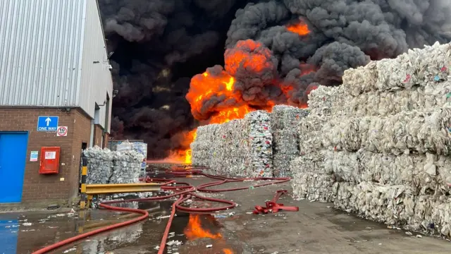 Flames and a black plume of smoke are shown rising from a recycling facility. In the foreground, a warehouse building is shown alongside bales of what appears to be plastic-based material.