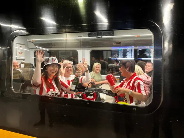 Waving Sunderland AFC fans, wearing red and white striped kit, sitting on a train. There is a group of four women - three of them wearing Sunderland colours. A woman in a hat nearest the window, on the left, holds a bottle of champagne.