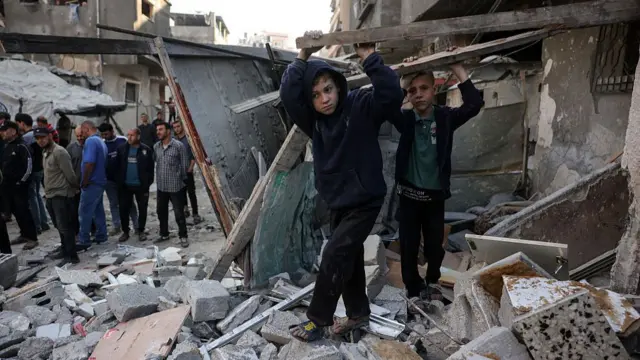 Two children stand on the rubbles of a bombed out home with their hands holding up wooden beams. A group of men stand on the left of the frame observing the damage