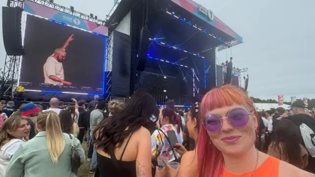 Romaine, wearing purple sunglasses, smiles at the camera in the crowd during the performance.