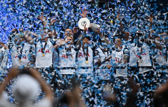The Tottenham Hotspurs players lift up the Europa Cup to cheers from fans, with blue confetti falling over them all.