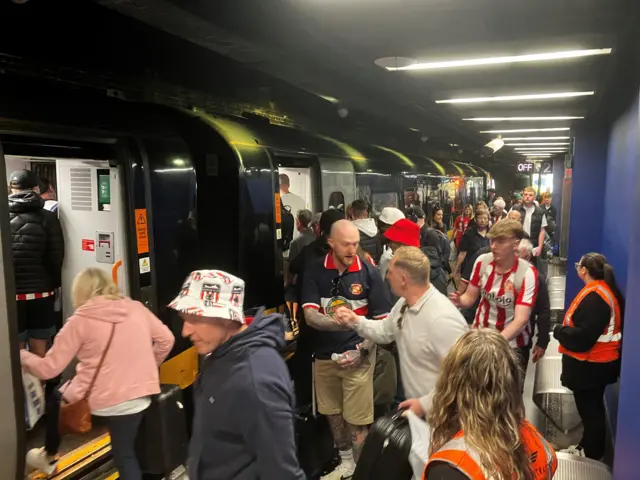 Sunderland AFC fans, many wearing red and white-striped kit, line up to board a train on a busy station platform in Sunderland.