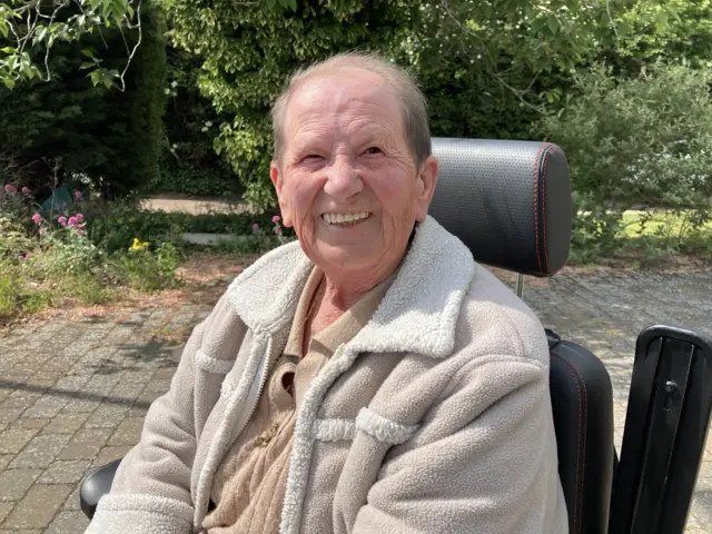 An elderly lady in a chair. She is wearing a sheepskin style fleece.