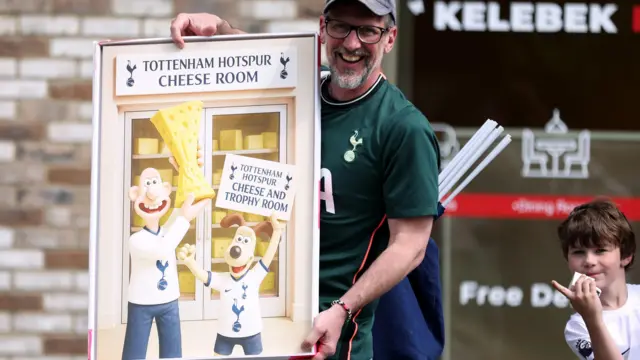 A man holds a poster of cartoon characters Wallace and Gromit. The characters are seen holding a cheese trophy cheering in front of a "cheese and trophy room"