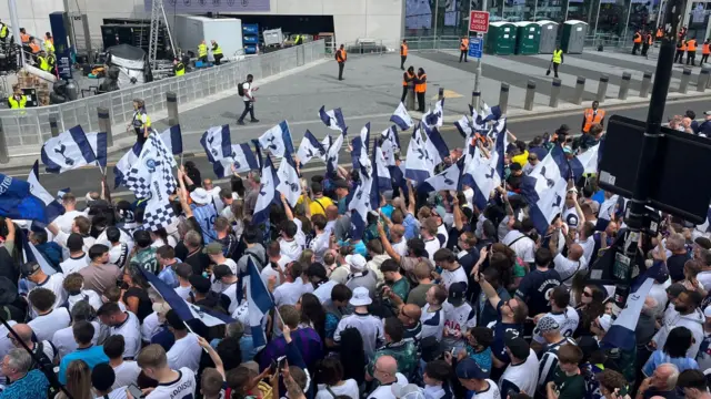 Fans lining the parade route near the stage lift their blue and white flags