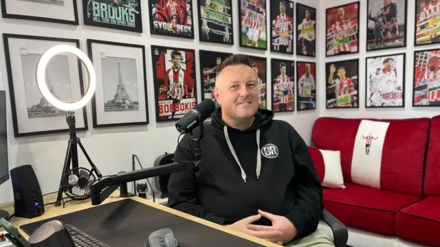 A man with short hair and wearing a dark hoodie sat in a recording studio. A mic is close to his face. Behind him is a red sofa and posters of footballers on the walls, as well as some pictures of the Eiffel Tower.