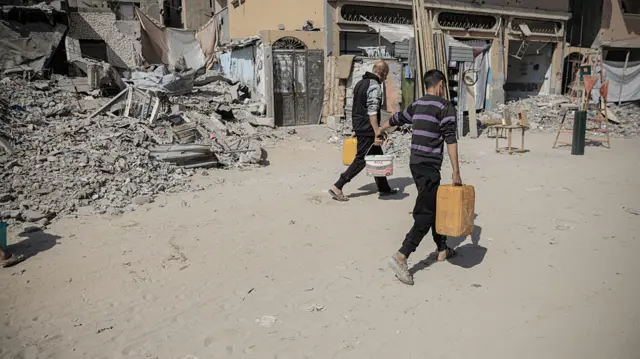 Two men carry cannisters as they walk through a street lined with rubble in Gaza
