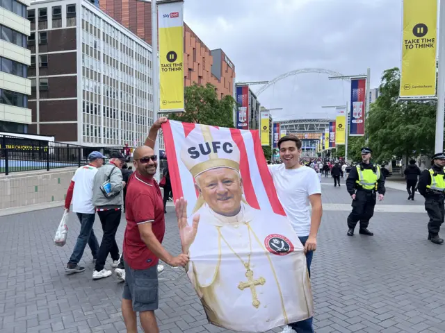 Two men hold up a flag depicting Chris Wilder as the Pope. The letters SUFC are emblazoned on his mitre