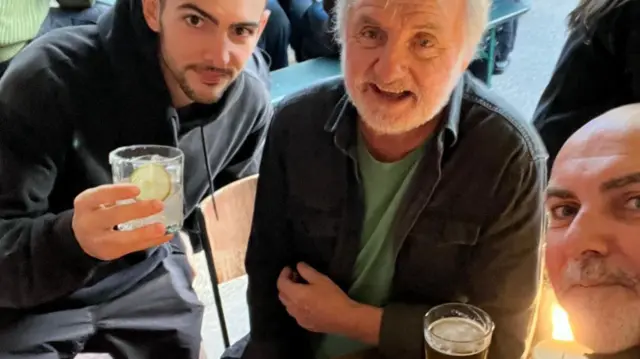 Three men sat in a pub. The one on the left is in his twenties and holding his drink aloft. The man in the middle is in his seventies and has a pint of beer next to his left arm on the table. The man on the right, who is taking the photo, is in his fifties.