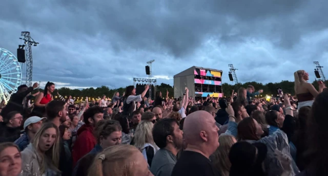 A crowd with arms up towards stage, with grey clouds above them.