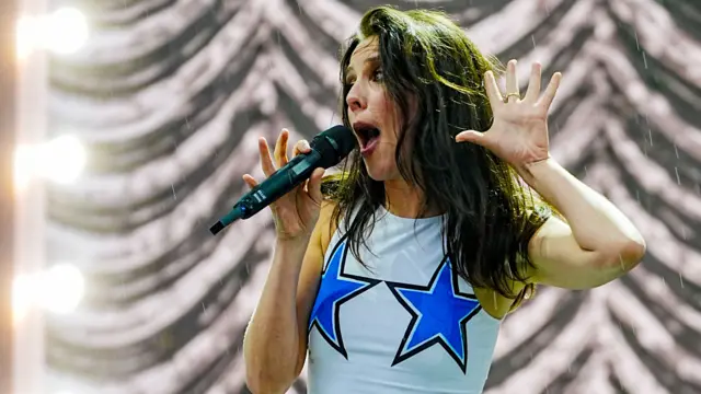 Ellie Rowsell sings into a microphone while holding a hand up. She has long brown hair and is wearing a sleeveless top with two blue stars on it.