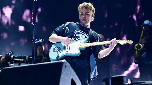 Sam Fender plays a light blue guitar on stage. He is wearing all black. He has brown hair.