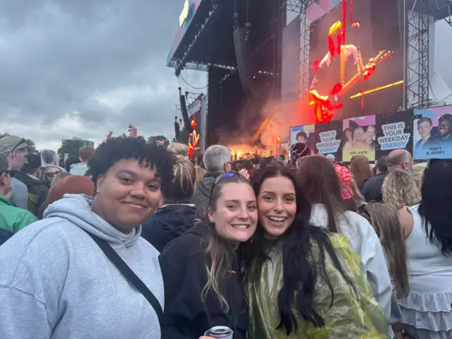 Taja, Hannah, and Mia smile for the camera while Sam Fender performs on the Main Stage behind them. They all have brown hair. Taja is wearing a grey jumper, Hannah is wearing a black jumper, and Mia is wearing a green poncho.