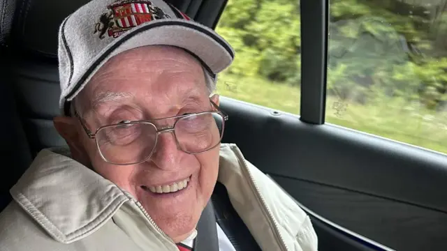 George Forester wears a Sunderland cap and a beige jacket. He is sitting in a car and is smiling.