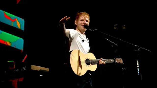 Ed Sheeran sings into a microphone on the New Music Stage at Big Weekend while holding a guitar. He is wearing a white t-shirt. He has ginger hair and blue eyes.