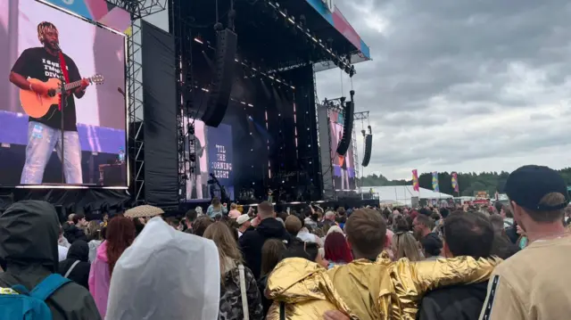 Crowds watch Myles Smith performing on stage with a guitar, with grey clouds overhead