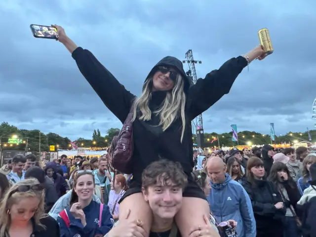 Jess sits on Henry's shoulders in a crowd at Big Weekend. Both are smiling at the camera. Jess has long blonde hair and Henry has curly brown hair.