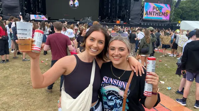 Katie and Katy smile for the camera while stood near a stage at Big Weekend. One of them has short brown hair and one has blonde hair. Both have blue eyes.