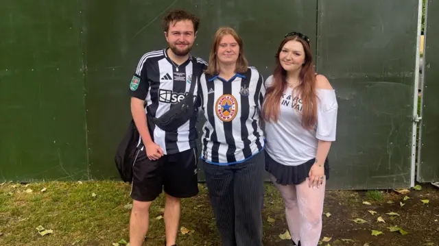 Three people stood in a line, they're wearing newcastle fc jerseys.
