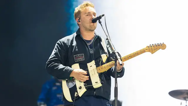 Sam Fender sings into a microphone while playing a guitar on stage at Big Weekend. He is wearing a navy jacket. He has brown hair.