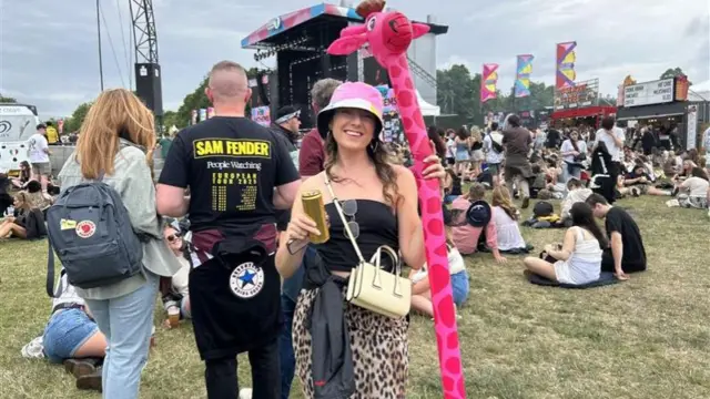 Alex Nelmes smiles for the camera in front of a stage at Big Weekend. She is holding a long-necked stuffed pink giraffe toy. Nelmes is wearing cheetah-printed trousers, a black top, and a pink bucket hat.