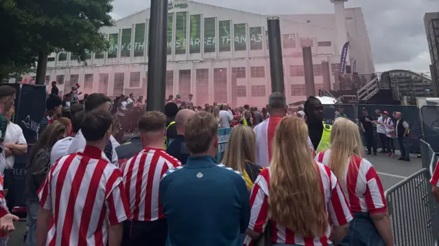 Sunderland fans wearing red and white striped kit wait at a Sunderland fan zone. There is red smoke and a large building in the background.