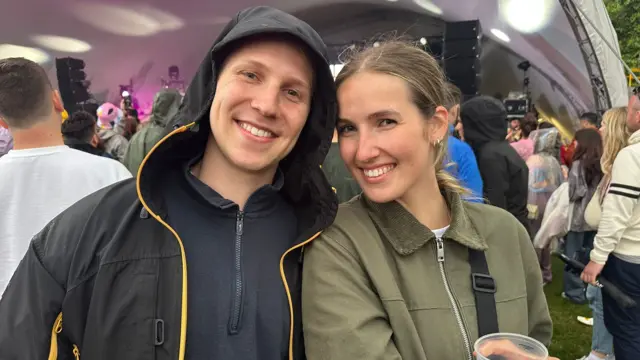 Monty and Amy smile for the camera while stood in a crowd in front of the BBC Introducing Stage. Monty is wearing a black coat with the hood up over his head and has blue eyes. Amy is wearing a khaki-coloured coat and has blue eyes and brown hair.