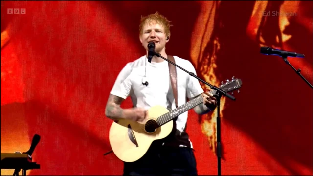 Ed Sheeran in white t-shirt and black trousers performs with acoustic guitar on stage at Big Weekend, a screen showcasing images in red and yellow behind him
