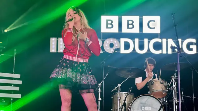 Luvcat sings into a microphone in front of a black and white BBC Introducing backdrop. She is wearing a red jacket and floral skirt. She has blonde and black hair.