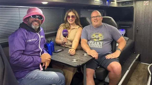 Kevin Duala and Claire Bryan sit on the Sugababes tour bus with its driver, Rob. They are all posing for the camera. Kevin is wearing a purple BBC Local Radio jacket, a pink bucket hat, and green sunglasses. Rob is wearing a grey graphic t-shirt. Claire is wearing a beige jumpsuit and brown sunglasses.