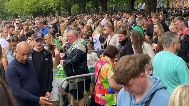 Large crowds queueing up outside Sefton Park in Liverpool. The groups vary in age and gender, at the forefront a teen in a light blue hoodie looking down