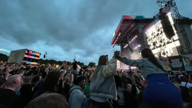 People sit on top of people's shoulders in a crowd in front of the Main Stage at Big Weekend that Sam Fender is performing on.