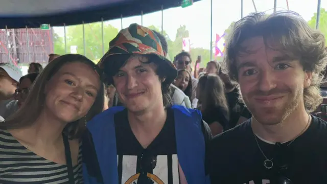 A long-haired blonde young woman (L) next to a man in a cap (C) and a bearded man (R) as they pose for pictures during a music show