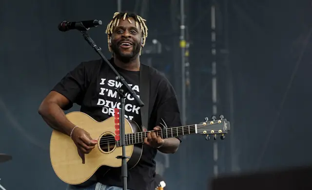 Myles Smith, wearing a black T-shirt and white writing, performs on the main stage with his guitar