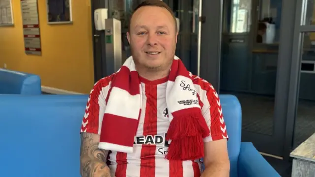 Andy Smith wears a Sunderland red and white striped shirt with a matching scarf. He sits on a blue sofa.