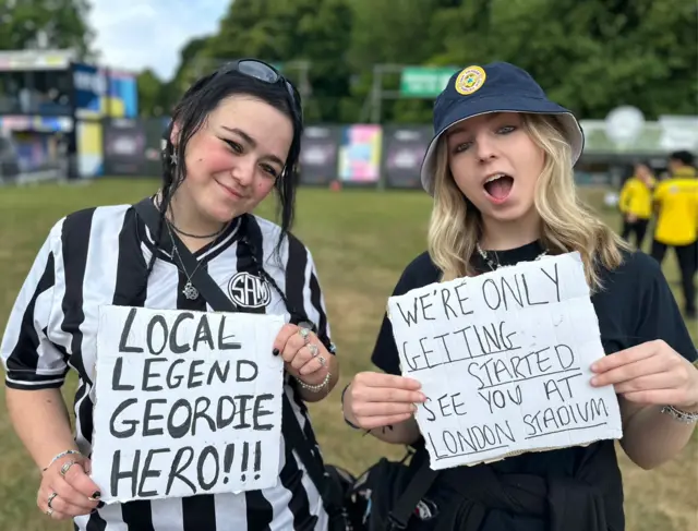 Chevy and Emily are dressed in black and white and hold up signs. One says Local legend Geordie Hero!!! and another that says We're only Getting Started See You at London Stadium.