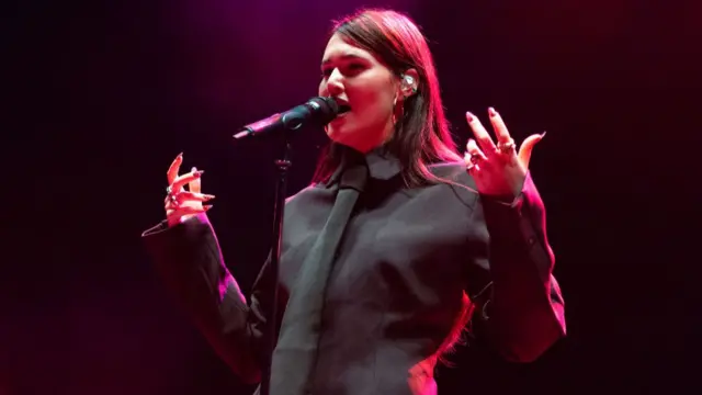 Sienna Spiro sings into a microphone. She is wearing a long-sleeves black shirt and a dark green tie. She has long brown hair.